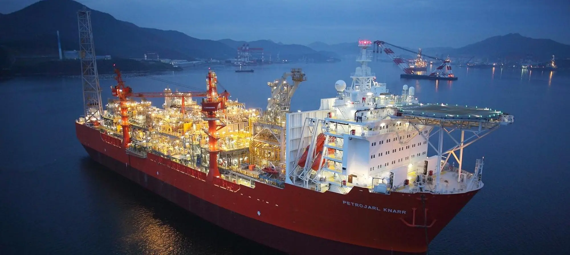 Large illuminated offshore production shipnamed Petrojarl Knarr docked in a harbour, with cranes and mountains visible in the background