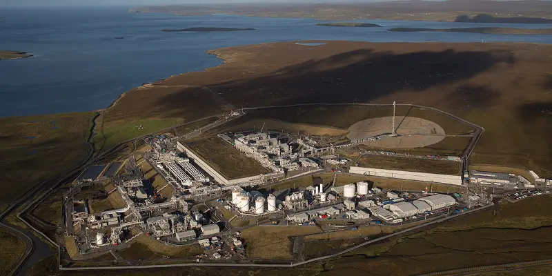 Aerial view of a large industrial complex with tanks and structures, surrounded by open land near a calm body of water